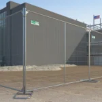 Construction site with metal safety fencing enclosing a modern building under construction; cranes and equipment visible in the background.