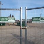 Pair of metal gates with Broadfence signs, closing off a fenced construction area on a paved lot under a blue sky