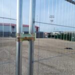 Close-up of a metal fence gate with a wooden spacer and padlock securing the gate; gravel lot and industrial buildings visible in the background.