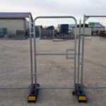 Portable metal gate on wheels standing in a gravel loading yard, with industrial buildings and equipment in the background.