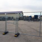 Temporary metal fencing with a closed gate enclosing a construction yard on a gravel lot, with a beige building and stacked materials in the background.