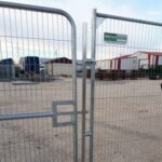 Metal security gate and fence blocking access to a construction yard with storage containers and portable buildings in the background.