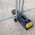 Portable metal barrier with a wheeled base and a yellow end cap, set on a dirt surface.