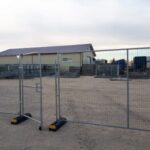 Construction site with metal temporary fencing and gates in a dirt lot, warehouse-style building and equipment visible in the background.