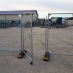 Two metal temporary fencing gates with base weights on a gravel lot outside an industrial yard.